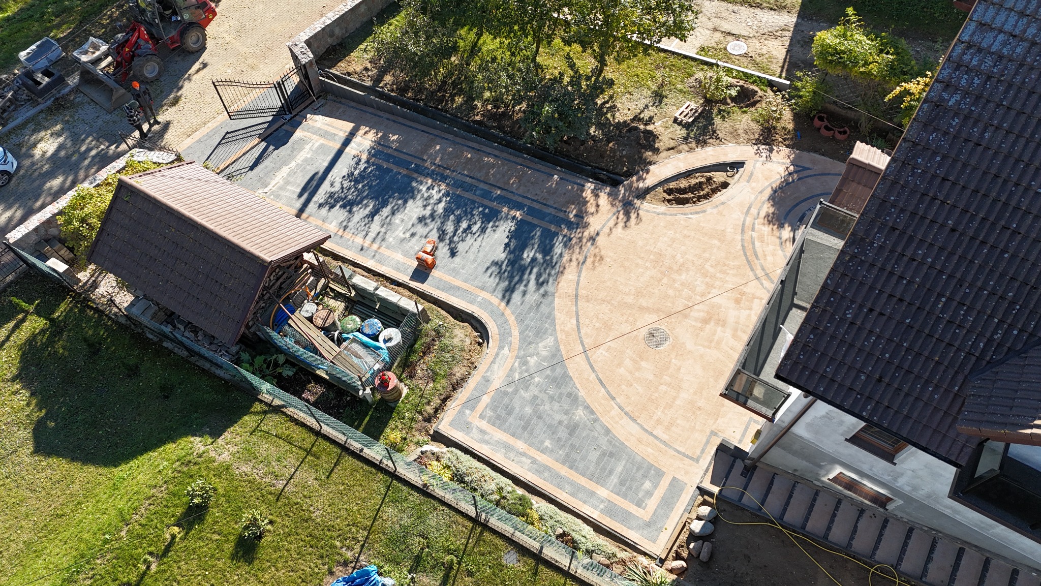 Widok z góry na posesję z nowo ułożoną kostką brukową w dwóch odcieniach, tworzącą wzór na podjeździe i ścieżkach, widoczny fragment budynku mieszkalnego, trawnik, ogrodzenie oraz sprzęt budowlany.