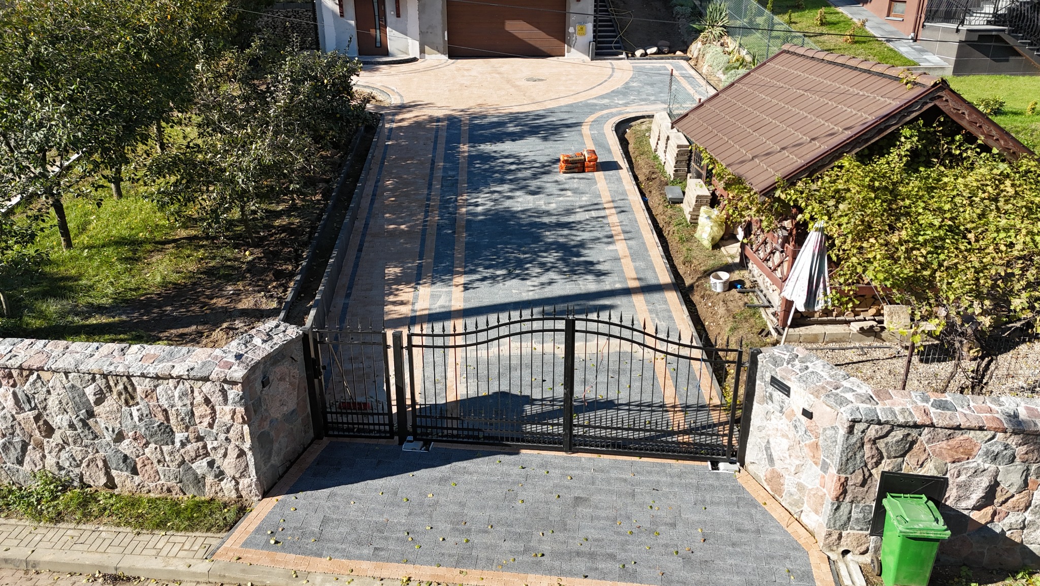 Widok z góry na posesję z bramą wjazdową, podjazdem z kostki w odcieniach szarości i beżu, oraz kamiennymi murkami po bokach, z widocznym fragmentem domu i ogrodu w tle.