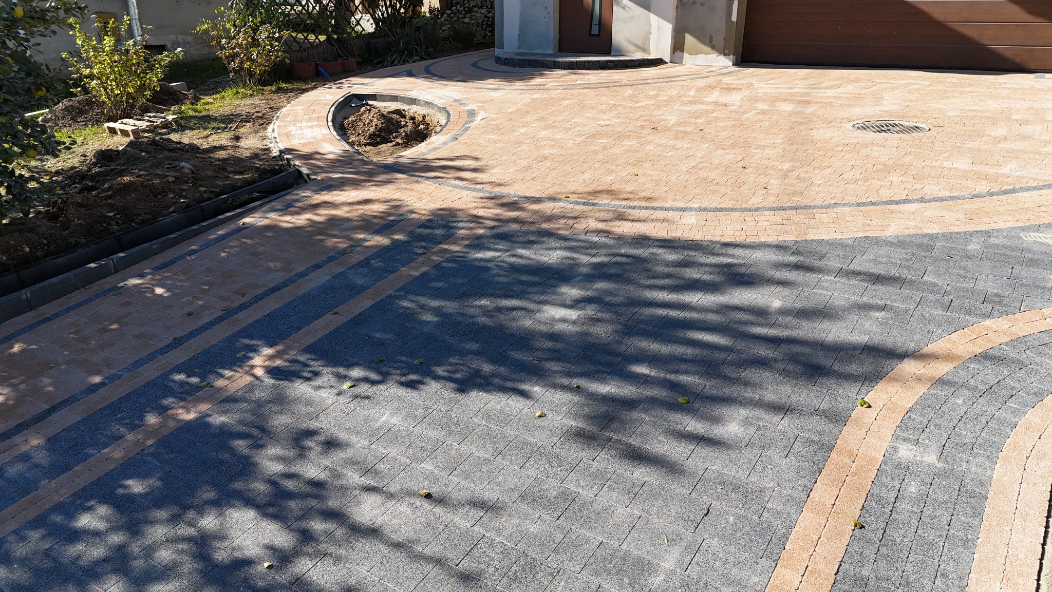Ułożona kostka brukowa w dwóch odcieniach szarości i beżu, z wykończeniem w formie okręgów i łuków, częściowo zacieniona przez drzewa, z widocznym przygotowanym miejscem pod nasadzenia roślin.