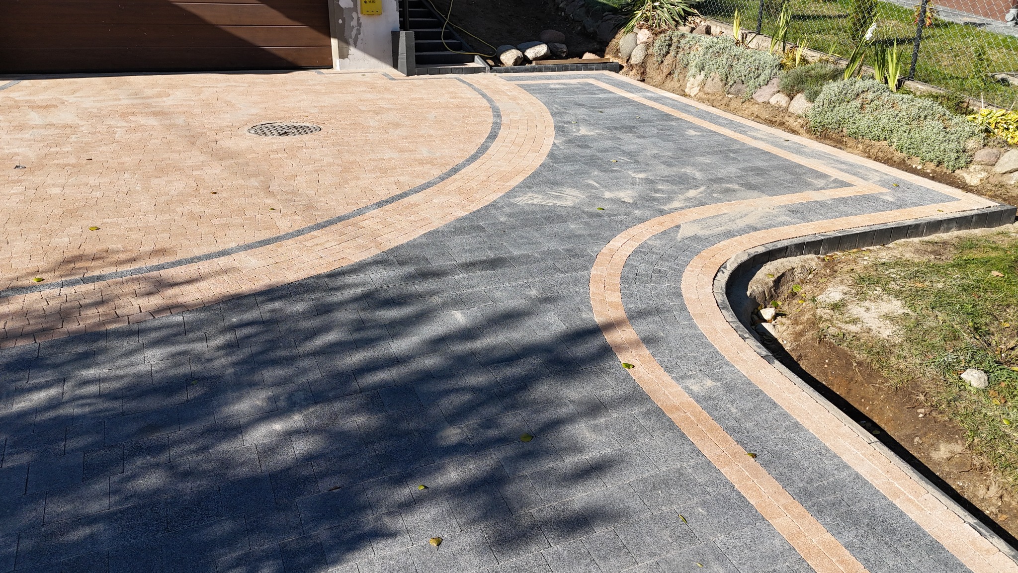 Podjazd z kostki brukowej w kolorach szarości i beżu, ułożony w łukowate wzory, z widoczną studzienką kanalizacyjną i obrzeżami, obok trawnik z roślinnością.