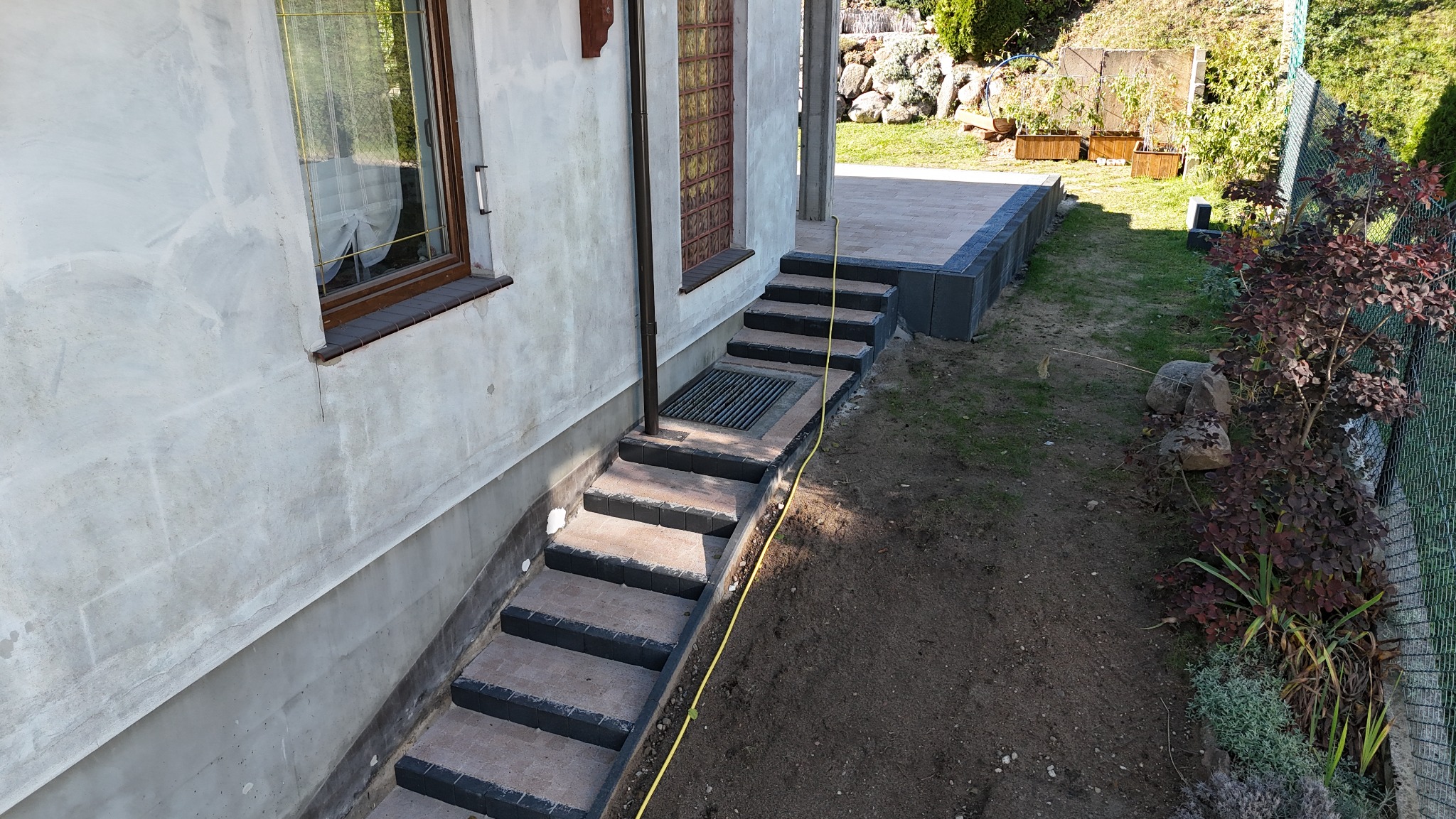 Zewnętrzne schody z kostki brukowej prowadzące do domu, z kratką ściekową i żółtym kablem leżącym obok, widok z góry na świeżo wyrównany teren.