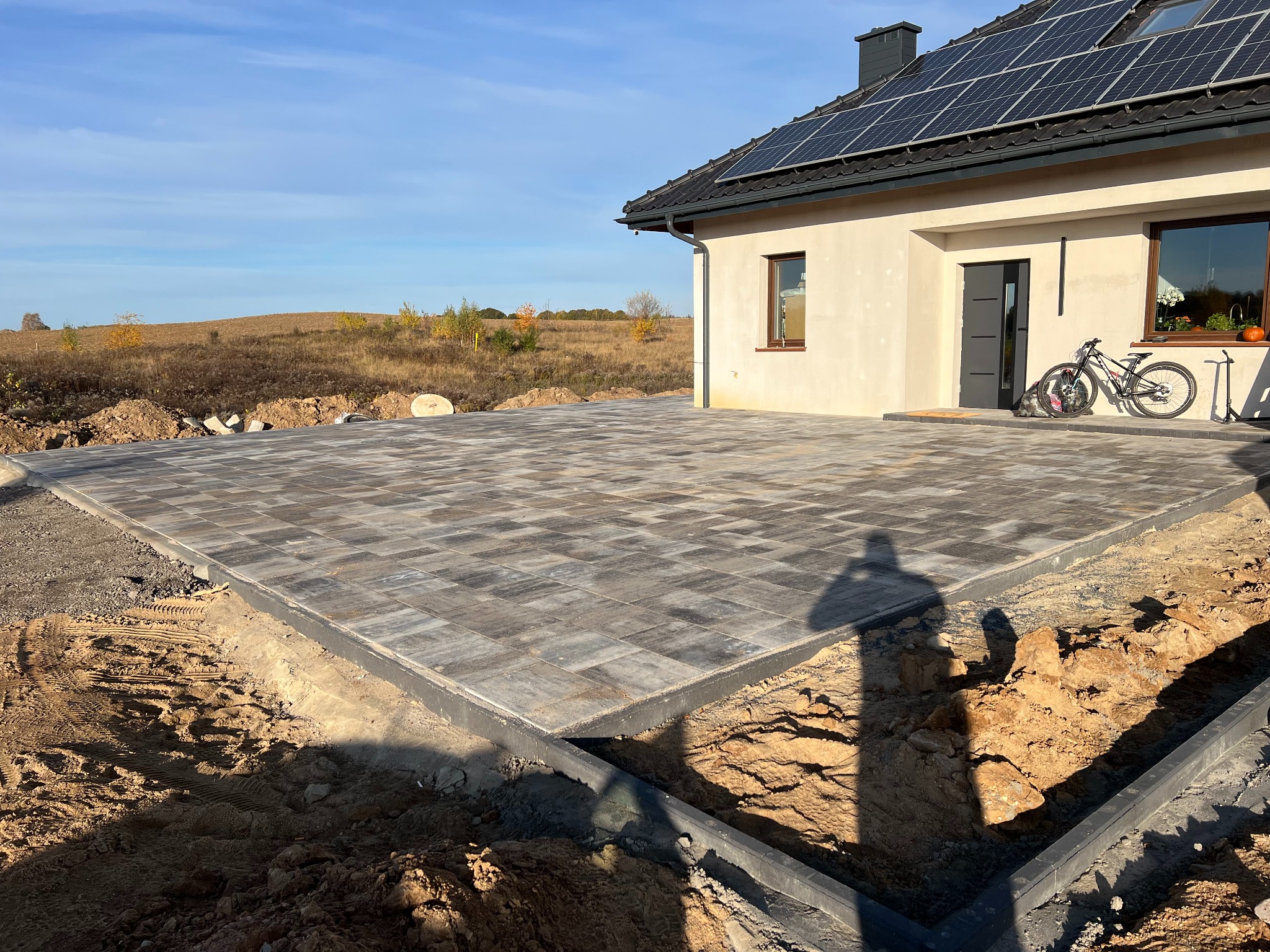 Nowo ułożona, szara kostka brukowa na posesji, z widocznymi obrzeżami i wykopem przygotowanym pod dalsze prace, dom z panelami słonecznymi w tle.