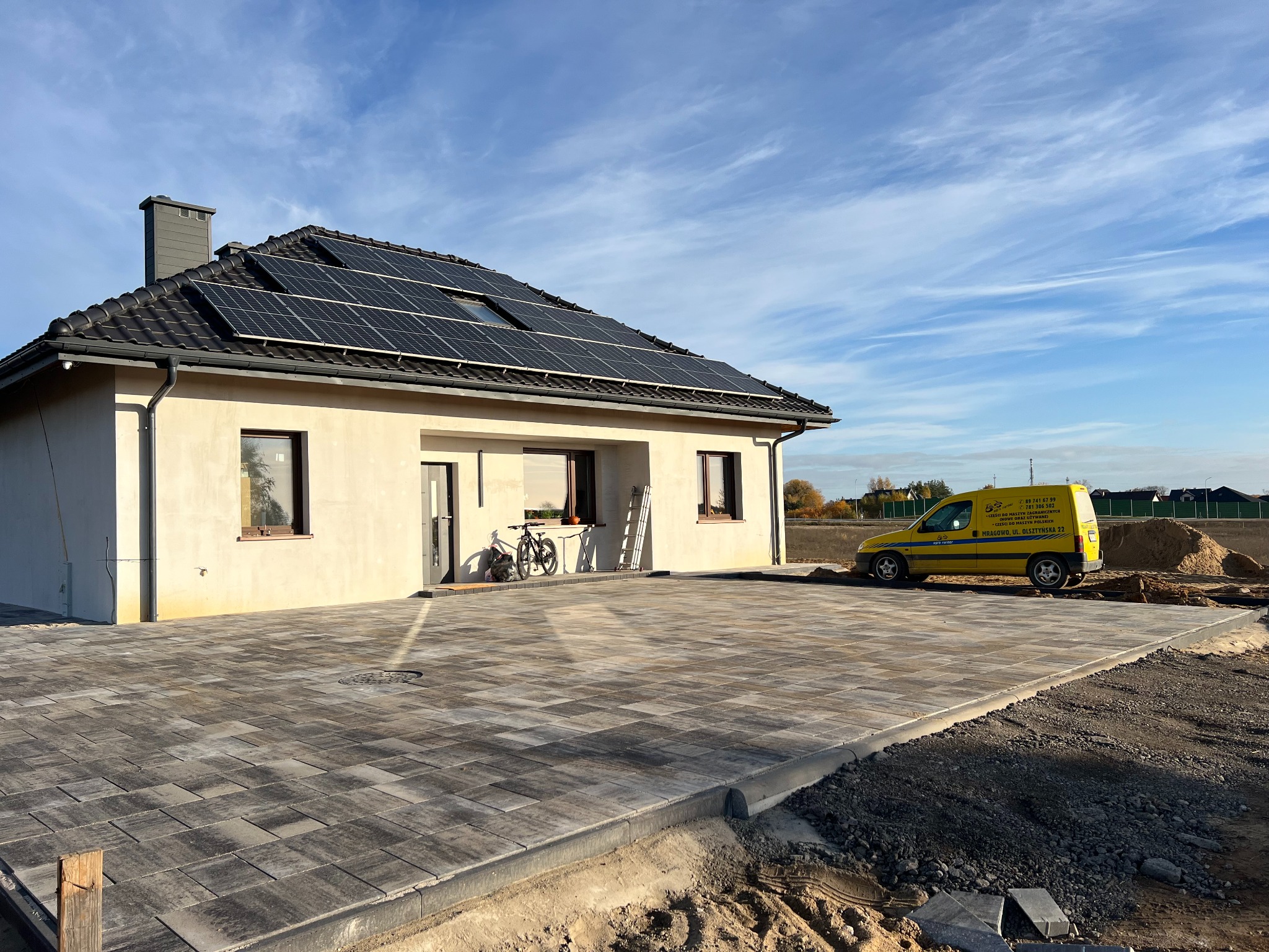 Nowo wybrukowany podjazd z szarej kostki przed domem z panelami słonecznymi, żółty samochód dostawczy z reklamą firmy w tle, rower oparty o ścianę budynku, w tle błękitne niebo