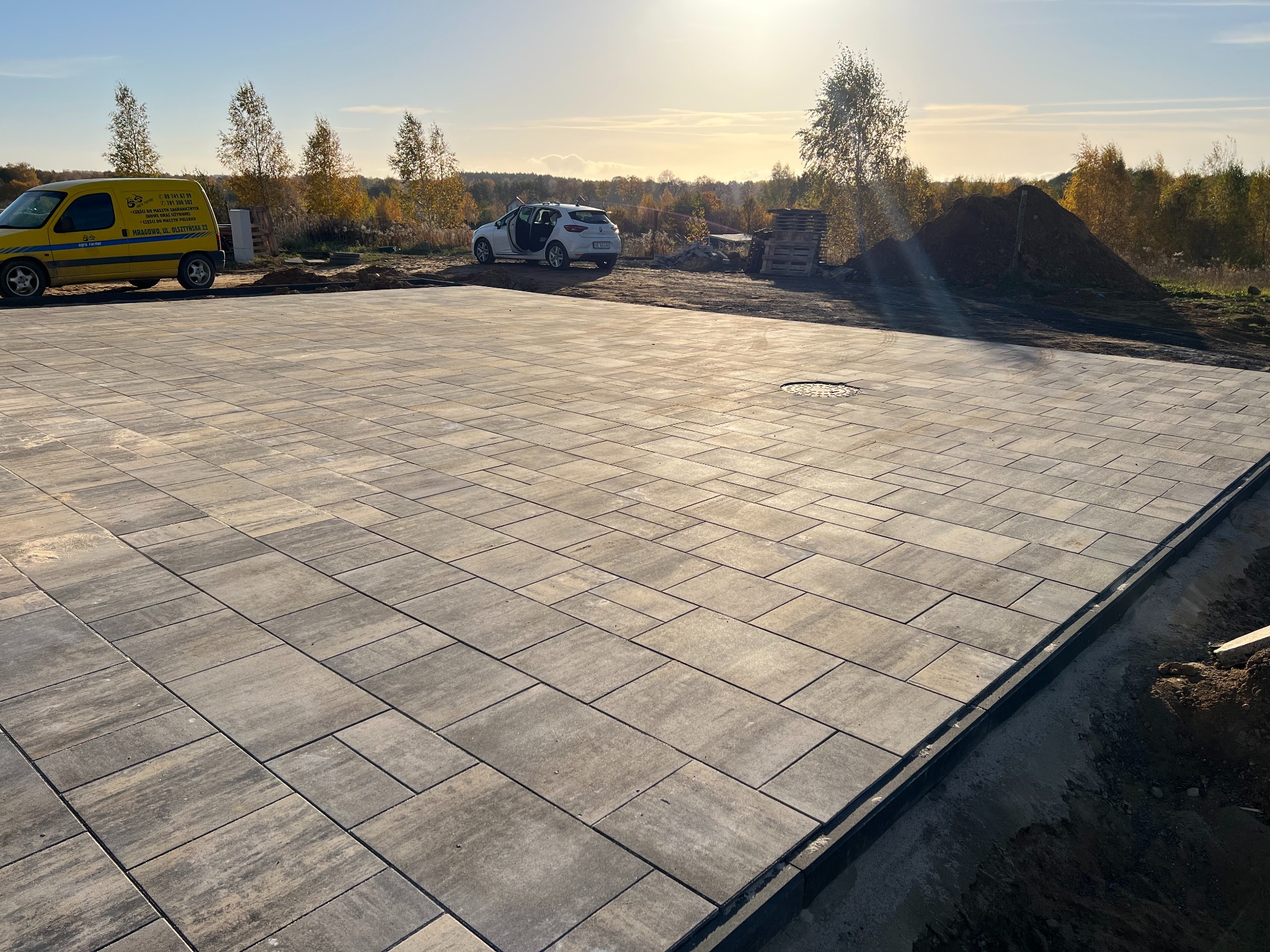 Ułożona nawierzchnia z szarych, prostokątnych płyt betonowych na posesji, z widocznym żółtym samochodem dostawczym z logo firmy, białym samochodem osobowym i stertą ziemi w tle, w słoneczny dzień.
