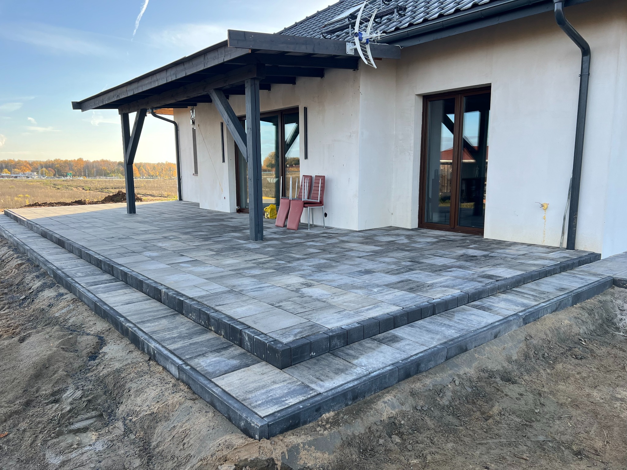 Nowoczesny taras z szarej kostki brukowej ułożonej w geometryczny wzór, z dwoma stopniami prowadzącymi do domu z białymi ścianami i ciemnym zadaszeniem. W tle widoczny krajobraz z drzewami...