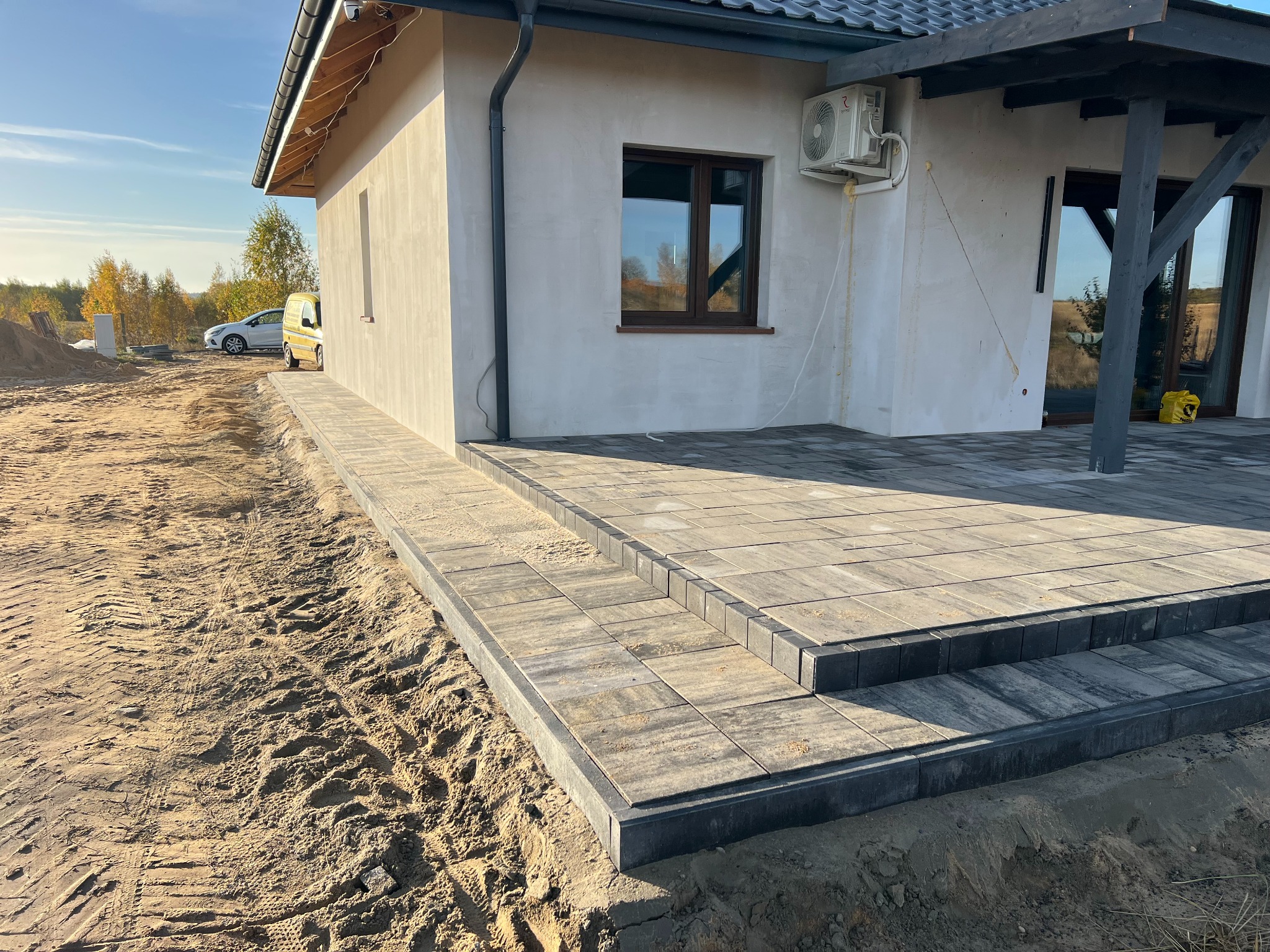 Ułożona kostka brukowa tworząca schody i chodnik przy nowym domu z klimatyzacją, widoczne ślady opon na piasku i samochód w tle.