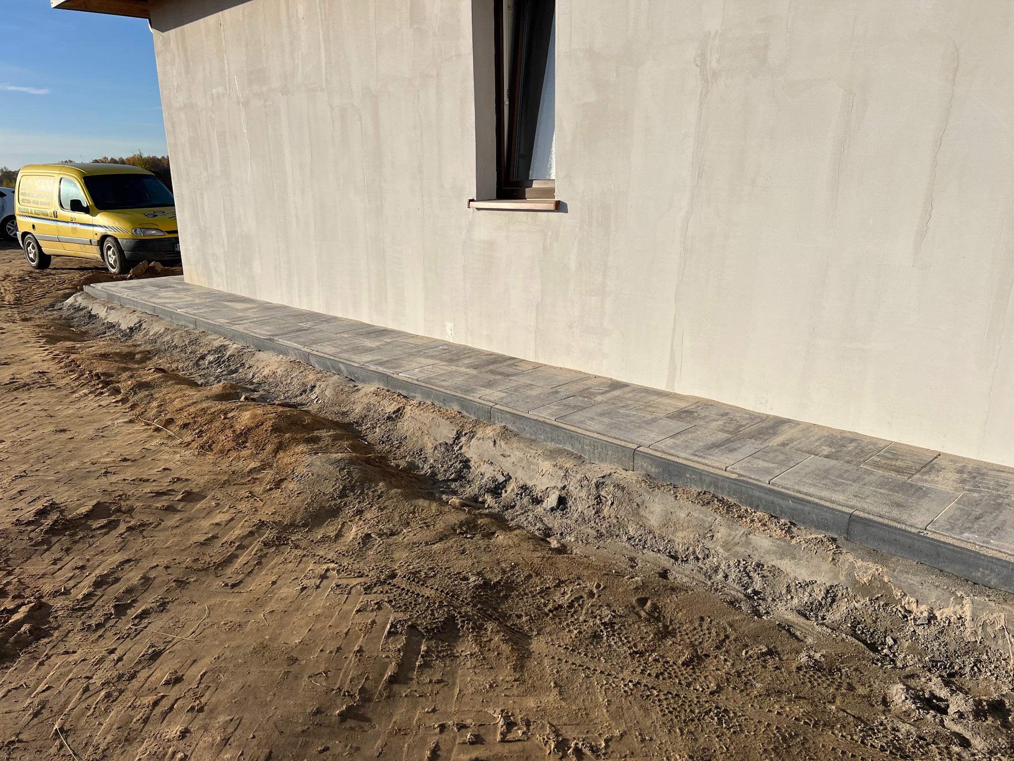 Świeżo ułożony chodnik z kostki brukowej przy ścianie budynku, z widoczną żółtą furgonetką w tle i nierównym terenem wokół.