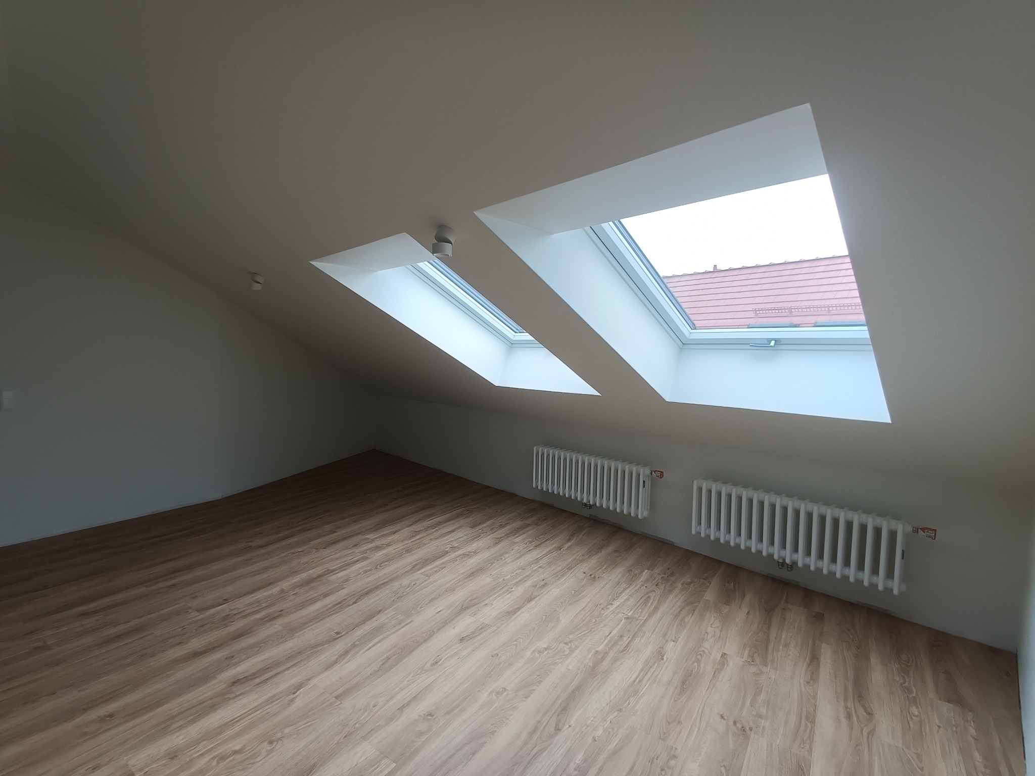 Poddasze z dwoma oknami dachowymi, świeżo ułożonymi panelami podłogowymi i białymi grzejnikami żeliwnymi pod oknami.