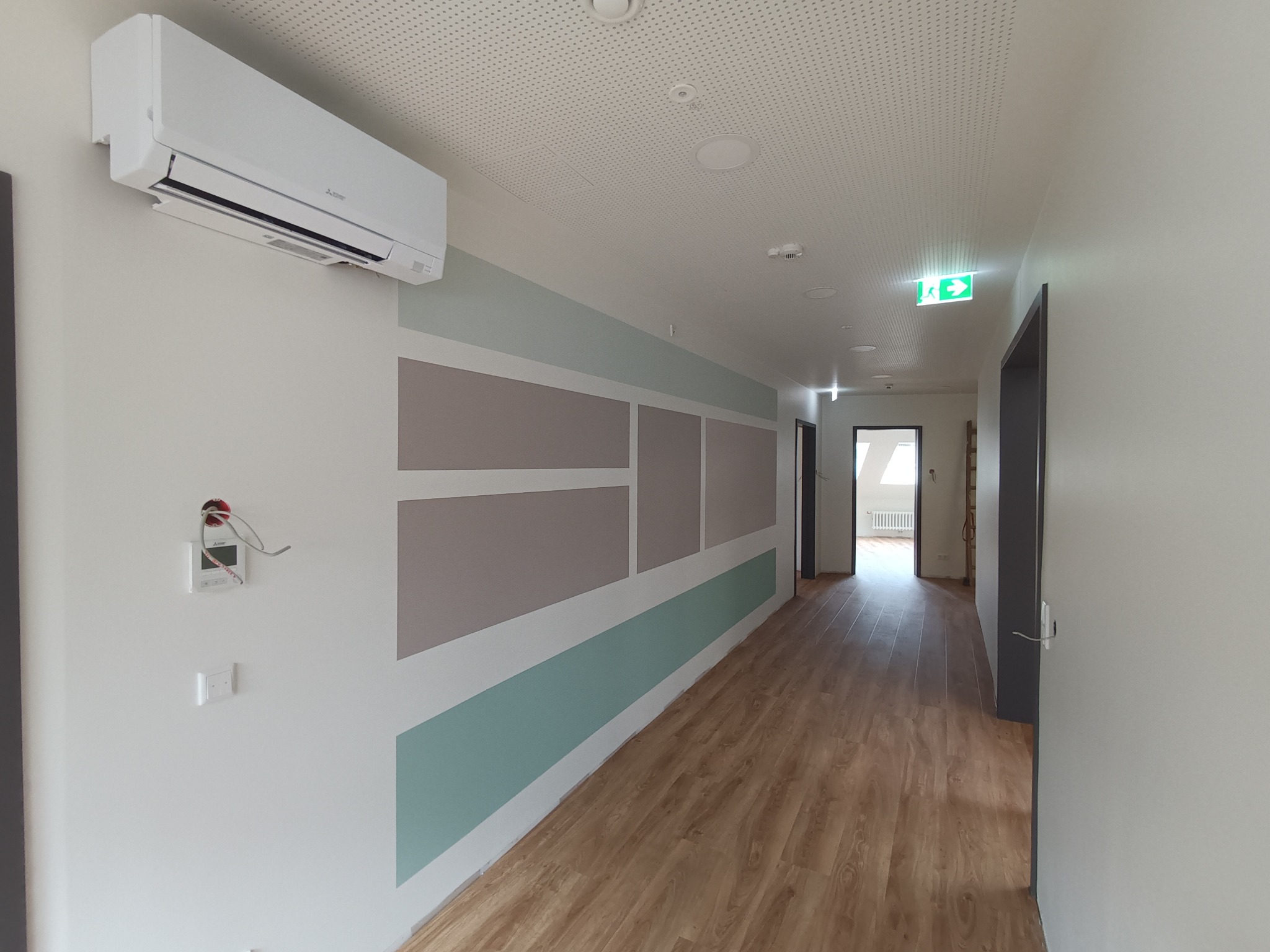Korytarz po remoncie z panelami podłogowymi, ścianami pomalowanymi w geometryczne wzory i zamontowanym klimatyzatorem Mitsubishi.