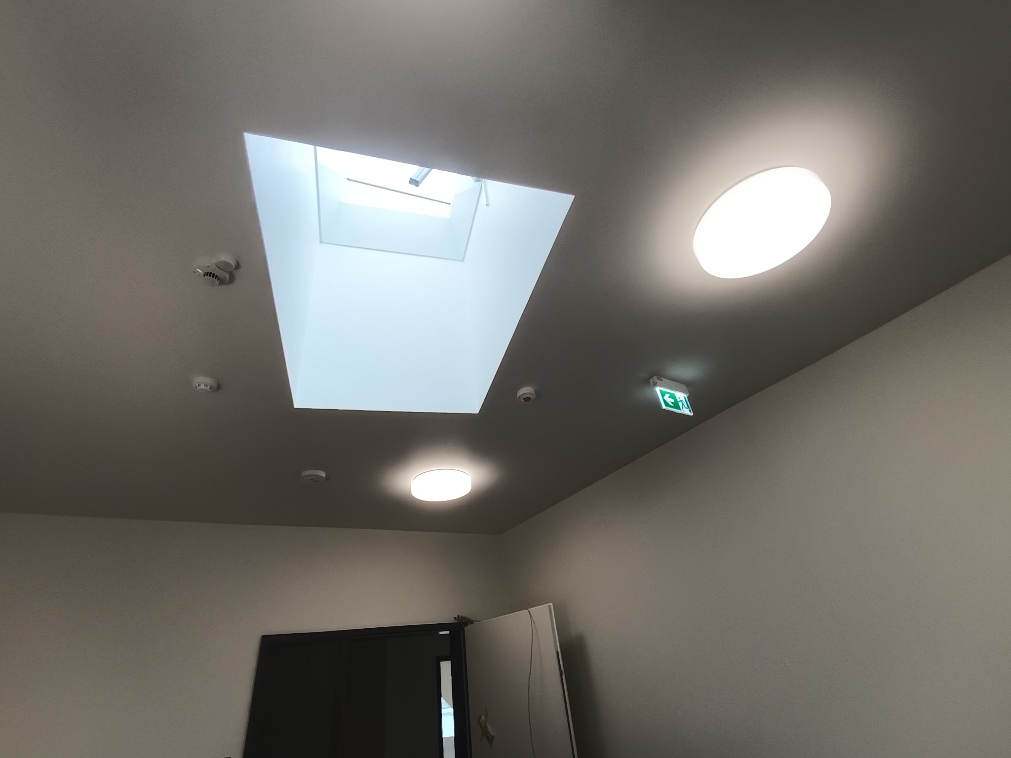 Szary sufit z kwadratowym świetlikiem, okrągłą lampą, czujnikami dymu i znakiem ewakuacyjnym