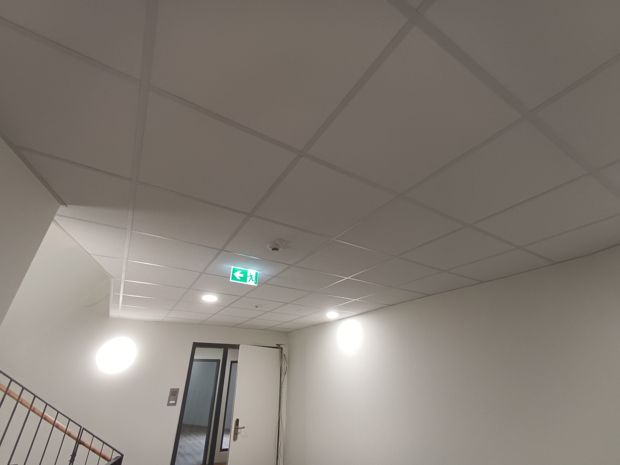 Sufit kasetonowy w biurowcu z oświetleniem punktowym i znakiem ewakuacyjnym, widok z dołu na klatce schodowej.