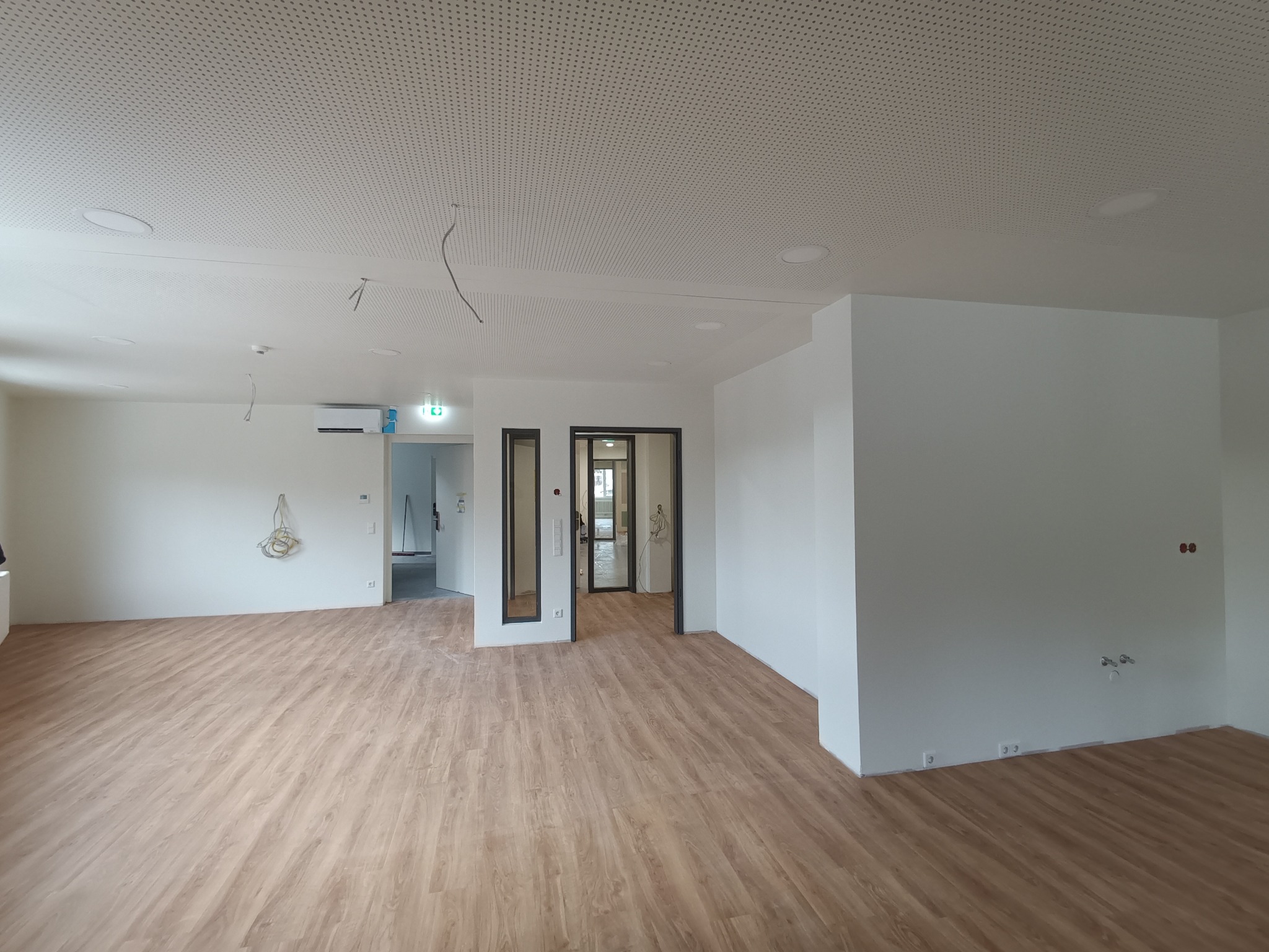 Wnętrze nowego pomieszczenia z jasnymi panelami podłogowymi, białymi ścianami i perforowanym sufitem akustycznym. Widoczne przygotowane punkty elektryczne i hydrauliczne na ścianie. Dwa otwarte...