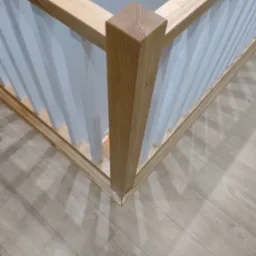 Fragment narożnika balustrady z jasnego drewna i białych pionowych elementów, widoczny na tle podłogi z paneli.