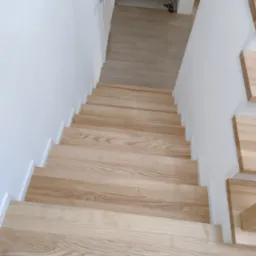Jasne, drewniane schody z białymi podstopnicami, widok z g&oacute;ry, minimalistyczny design, w tle drzwi i podłoga z paneli.