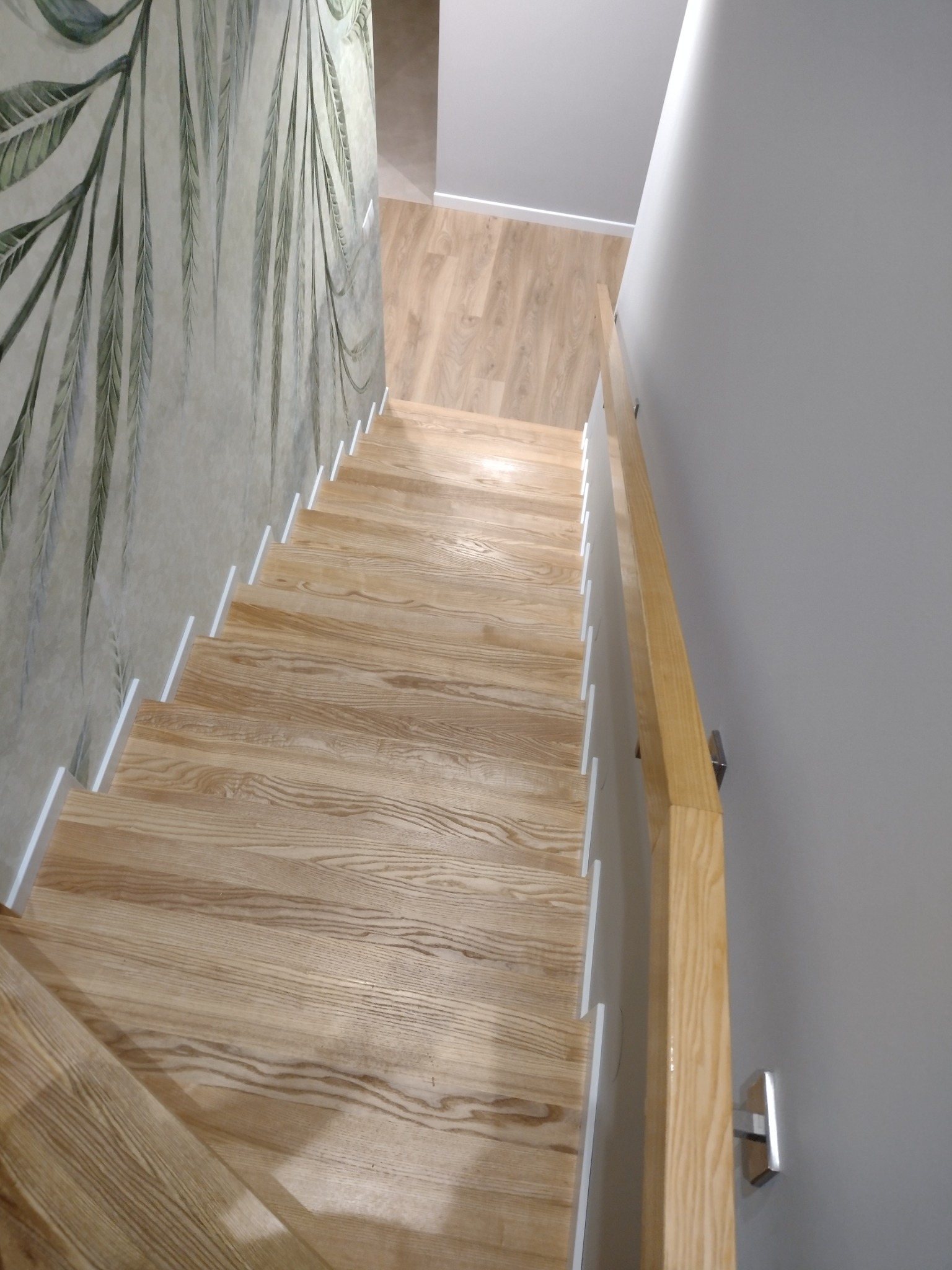Jasne, drewniane schody widziane z góry, z białą listwą przypodłogową i drewnianą poręczą zamontowaną na szarej ścianie, obok ściany z tapetą w motywy roślinne.