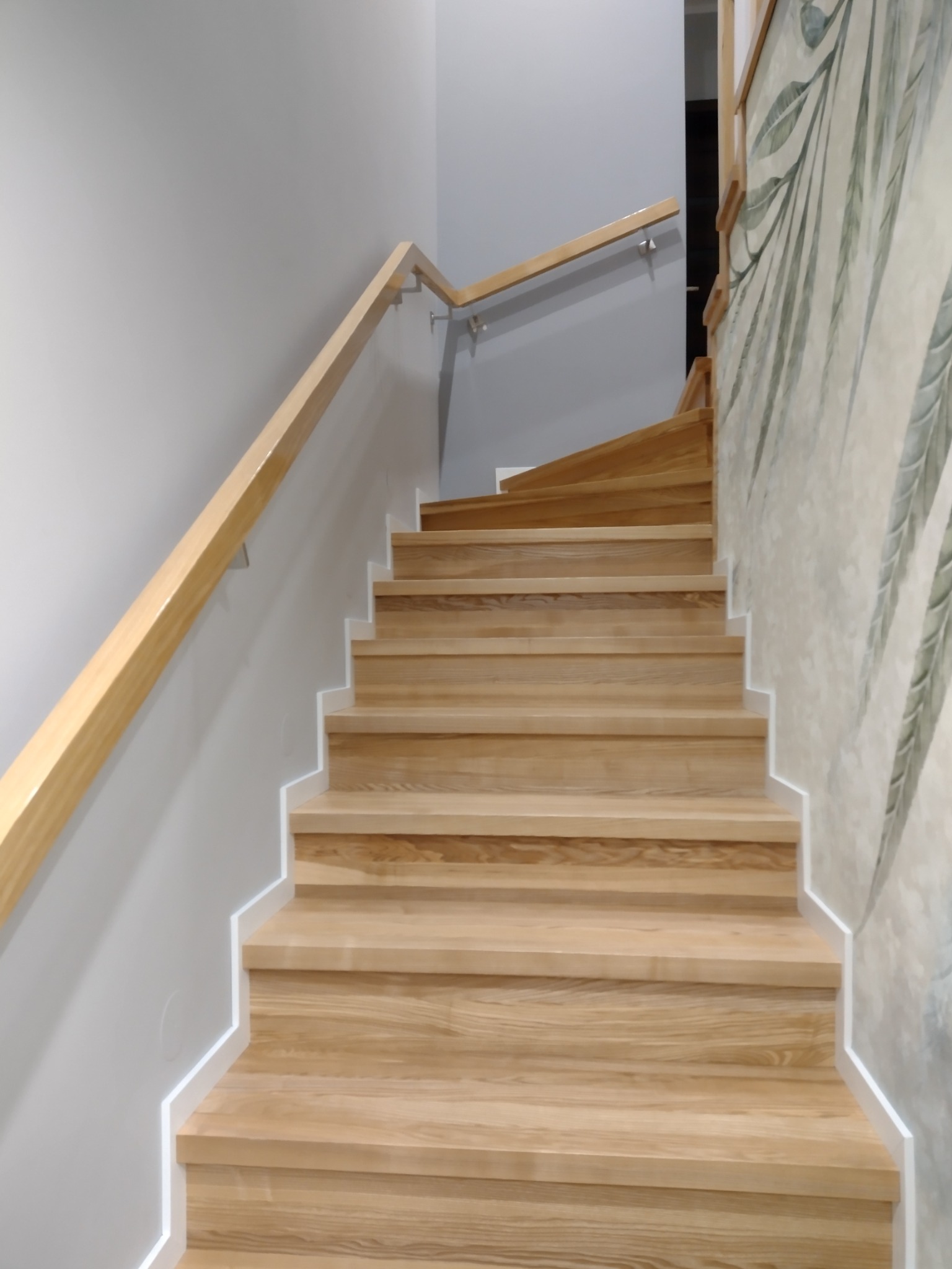 Jasne drewniane schody z poręczą zamontowane w nowoczesnym wnętrzu z szarą ścianą i tapetą w motywy roślinne.