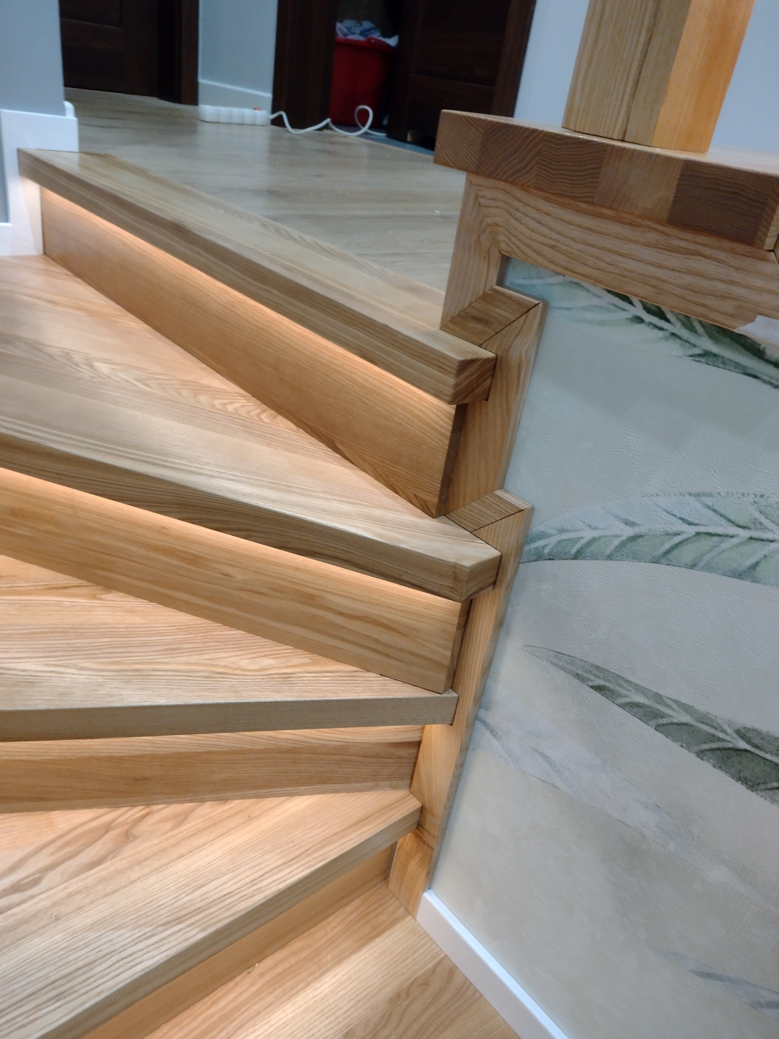 Drewniane schody z podświetleniem LED pod stopniami, widok z góry na zakręt, z fragmentem balustrady i wzorem liści na ścianie.