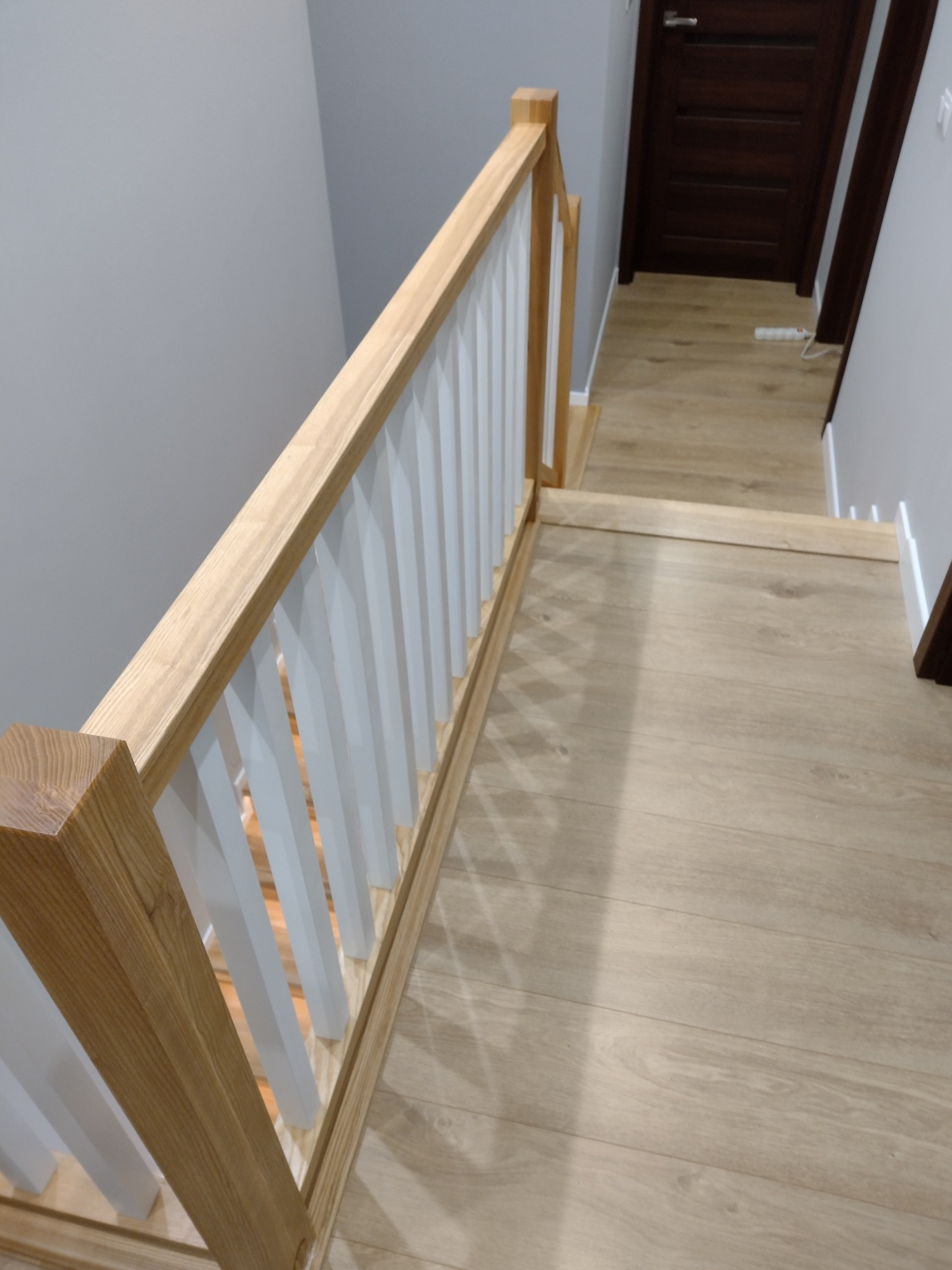 Jasne, drewniane schody z białą balustradą, widok z góry, parkiet na podłodze, ciemne drzwi w tle.