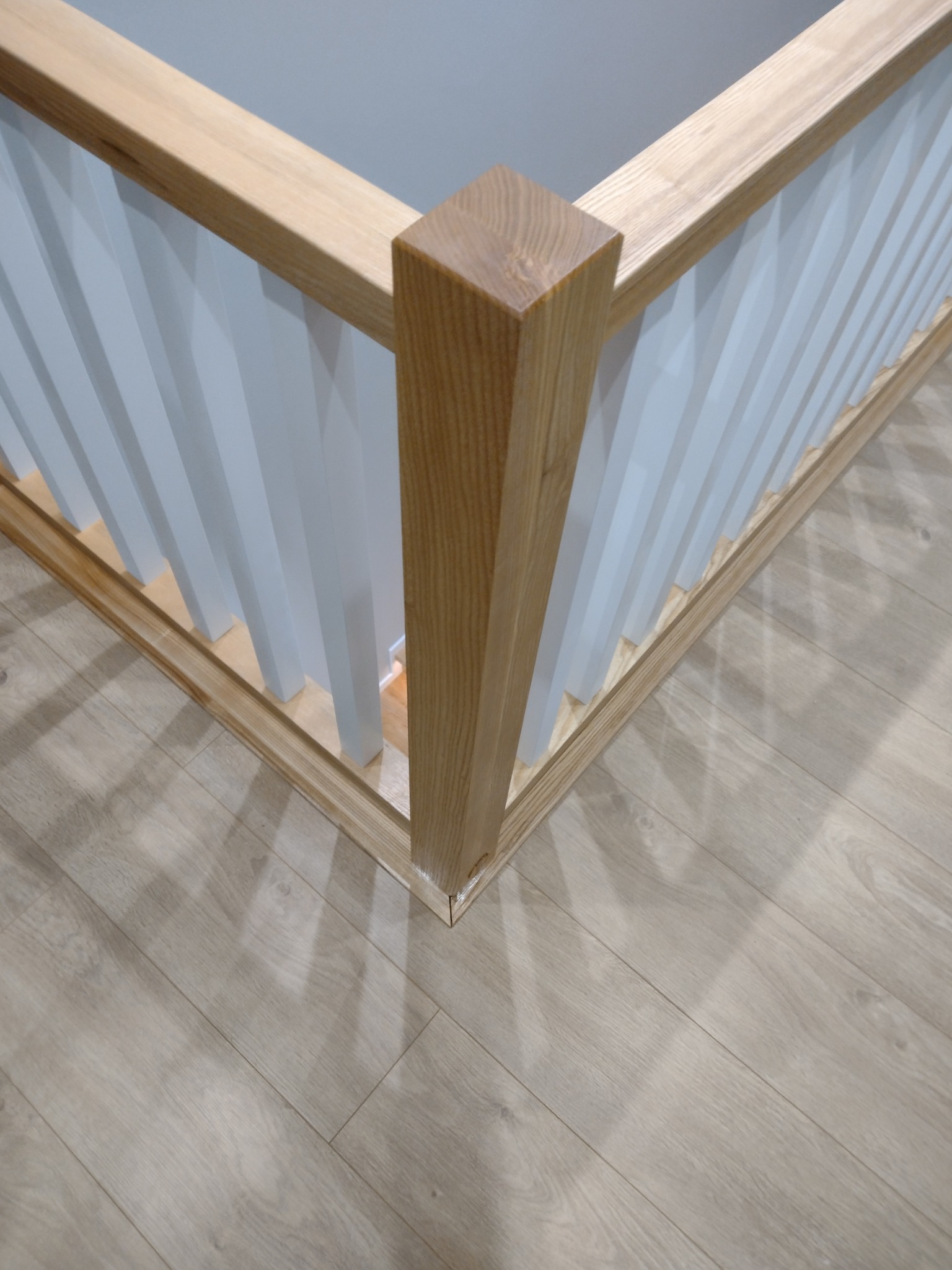 Fragment narożnika balustrady z jasnego drewna i białych pionowych elementów, widoczny na tle podłogi z paneli.