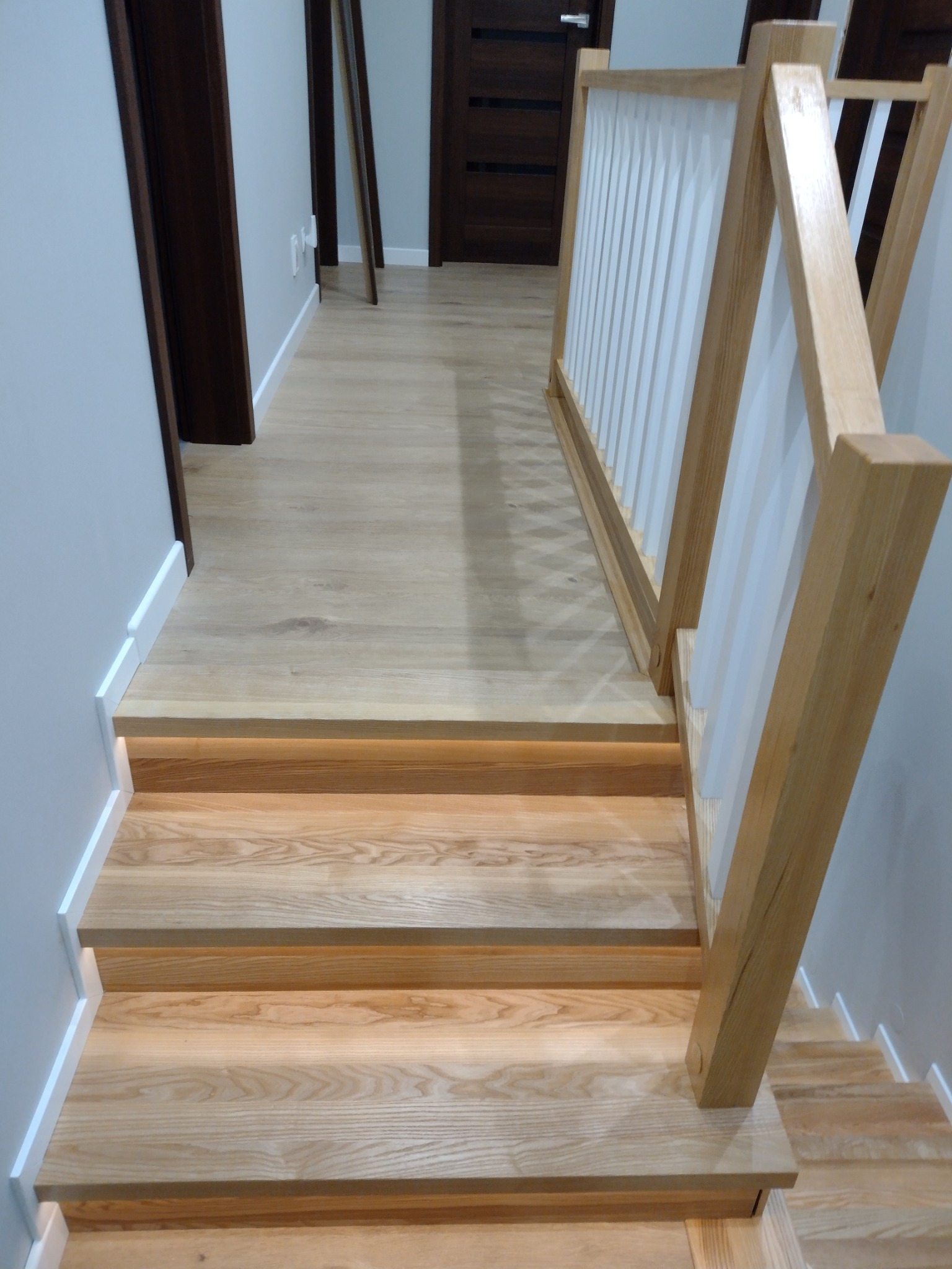 Jasne drewniane schody z podświetleniem LED pod stopniami i biało-drewnianą balustradą z pionowymi panelami, widok z góry na dół.