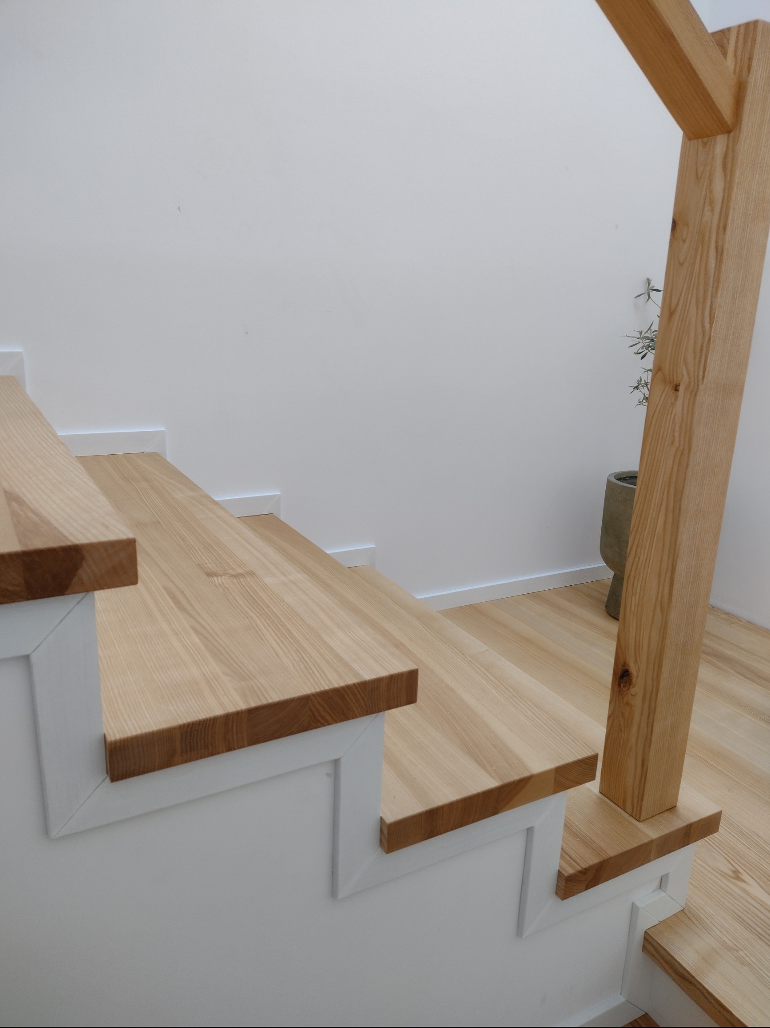 Jasne drewniane stopnie schodów z białymi podstopnicami, widoczny fragment drewnianej balustrady i podłogi, minimalistyczne wnętrze.