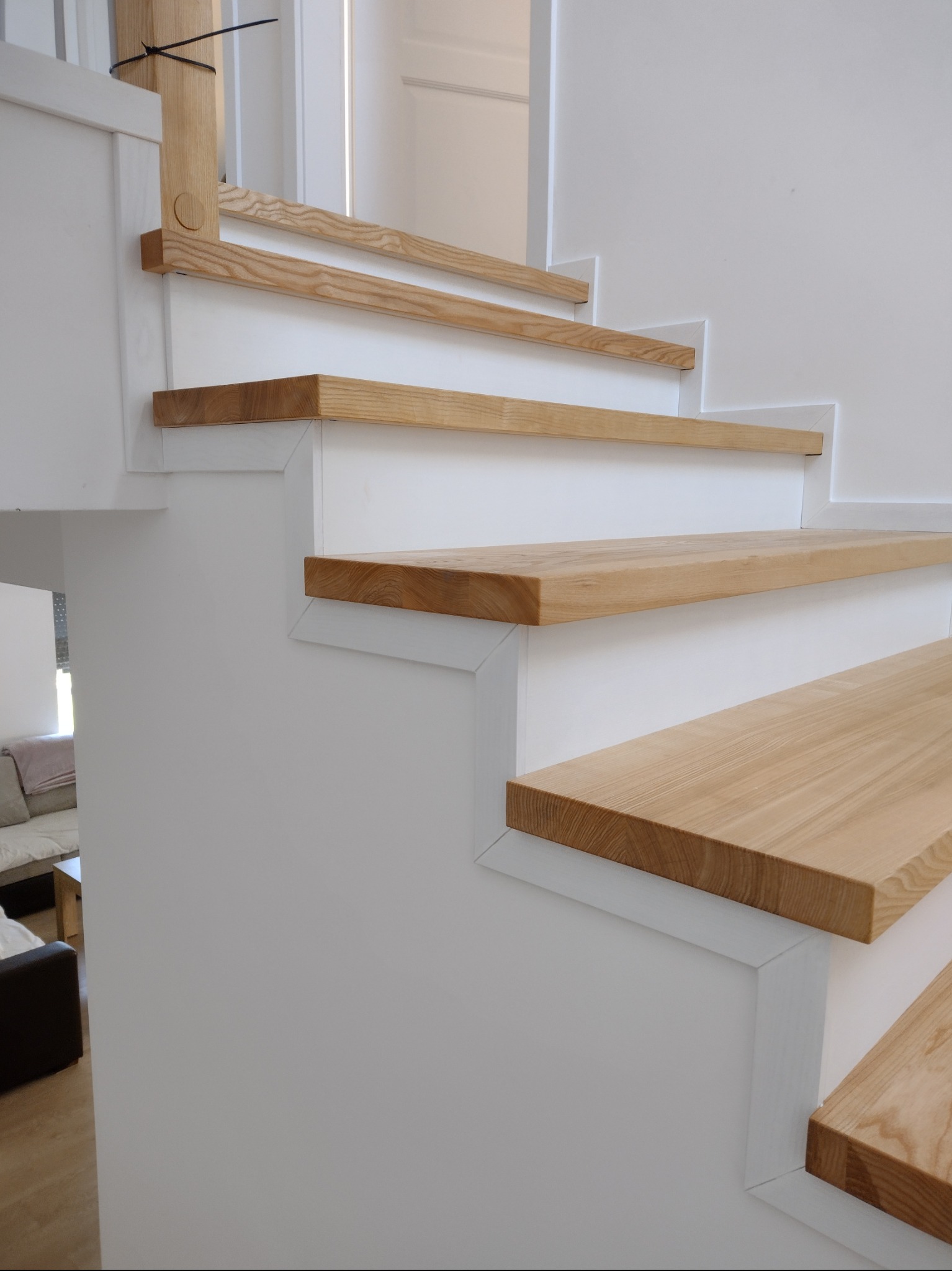 Nowoczesne schody z jasnego drewna i białych elementów konstrukcyjnych, widoczne z bliskiej perspektywy, z fragmentem salonu w tle.