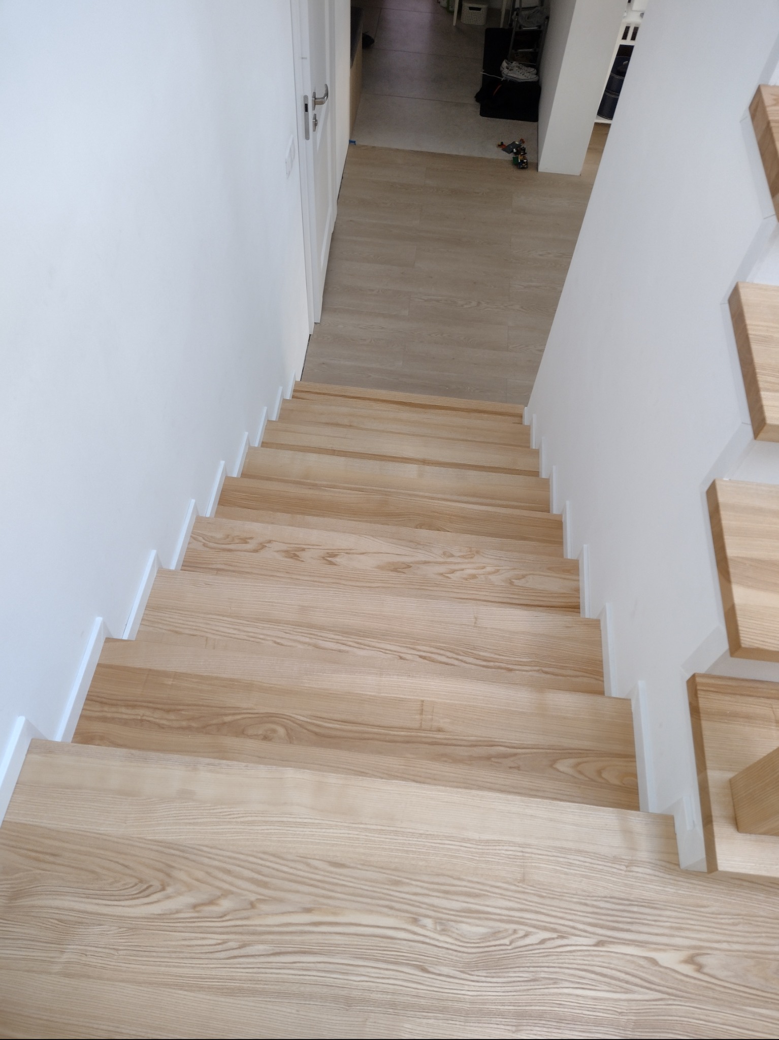 Jasne, drewniane schody z białymi podstopnicami, widok z góry, minimalistyczny design, w tle drzwi i podłoga z paneli.