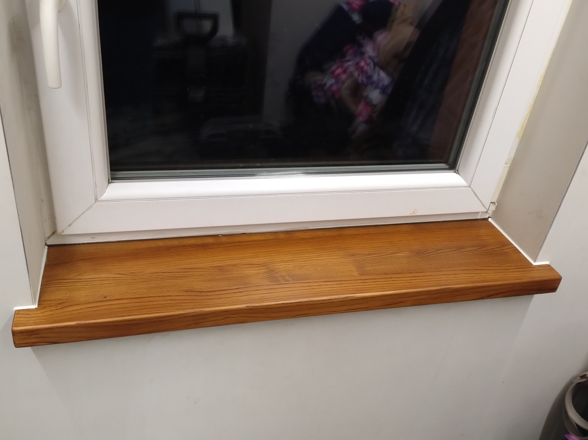 Zamontowany, brązowy parapet drewniany pod oknem PCV. Widoczne uszczelnienie silikonem. Ujęcie z dołu, podkreślające strukturę drewna i detale wykończenia.