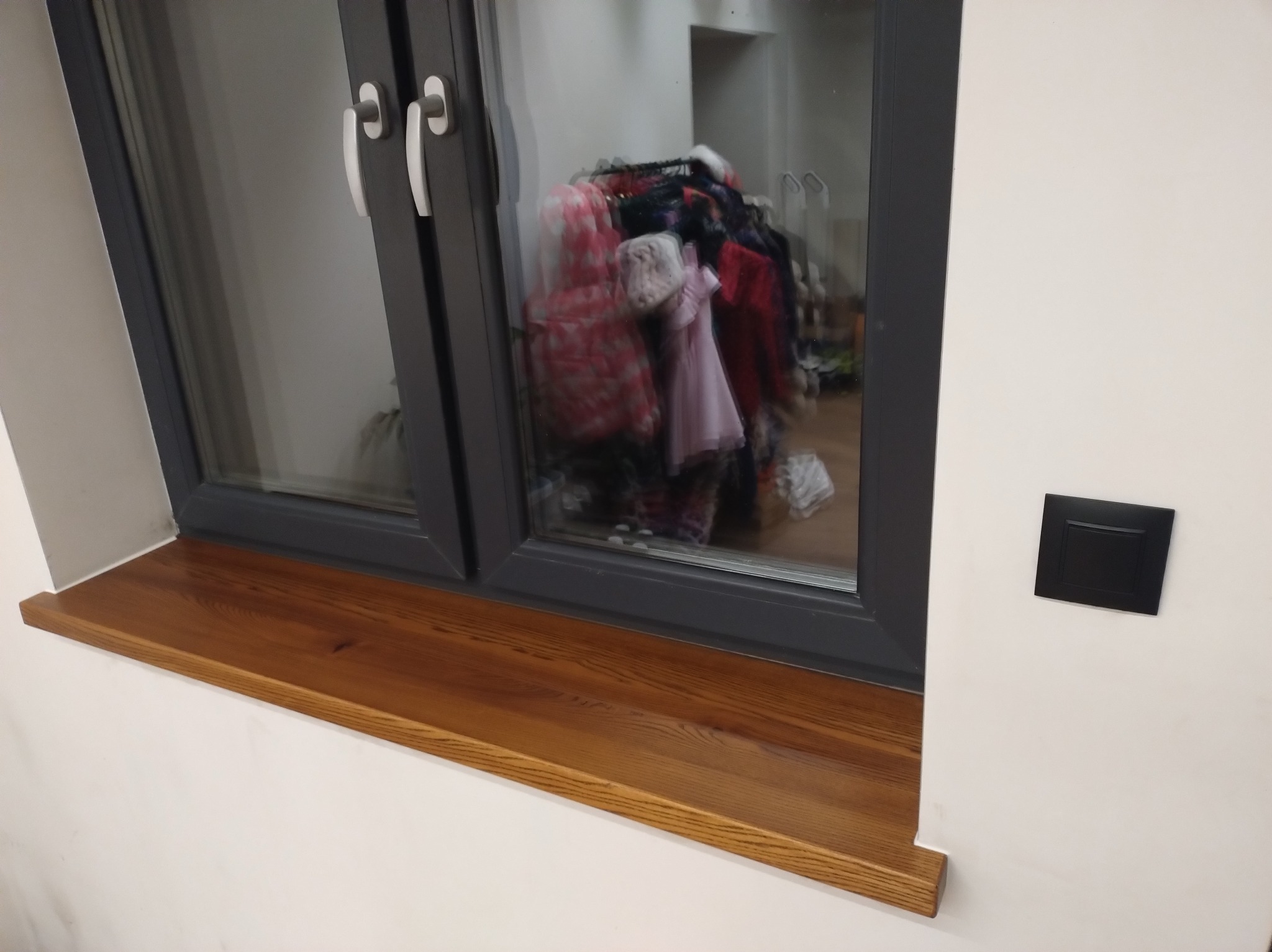 Zamontowany drewniany parapet o ciepłym odcieniu brązu pod oknem z szarą ramą. Widoczny włącznik światła na ścianie. W tle, za oknem, widoczne ubrania na wieszaku.