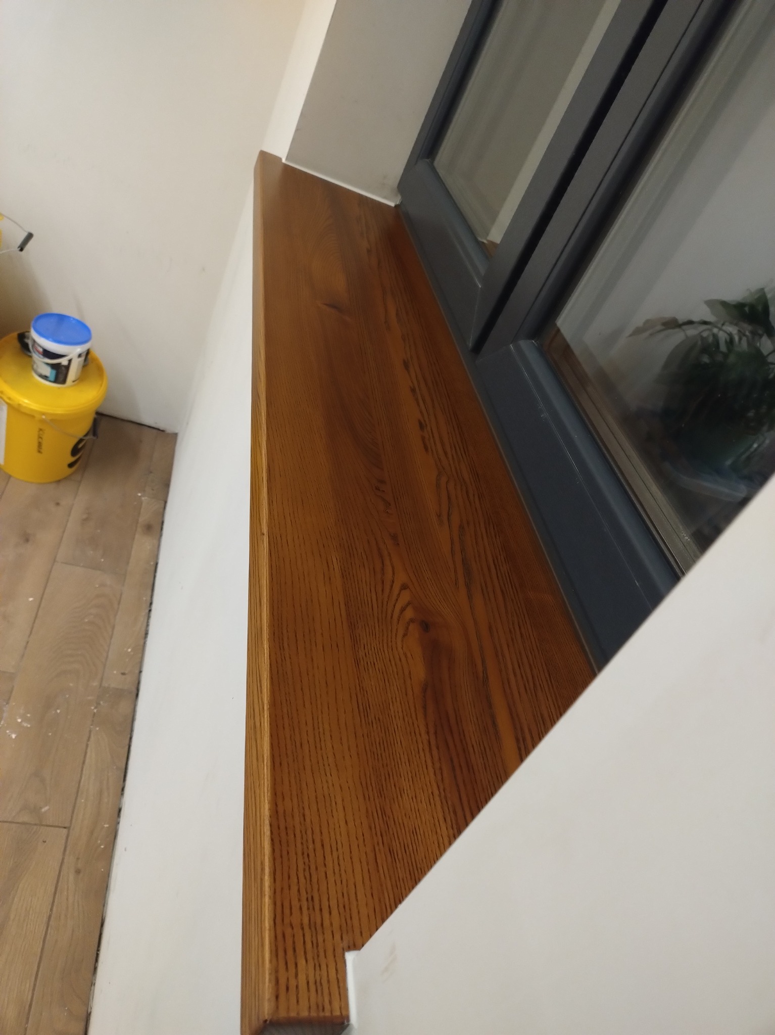 Zamontowany drewniany parapet o ciepłym odcieniu brązu, przylegający do okna z szarą ramą. Widoczny fragment podłogi i wiadro z farbą.