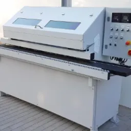 Profesjonalna, stacjonarna maszyna stolarska z panelem sterowania, widoczne elementy transportowe i osłony, ustawiona w jasnym pomieszczeniu.