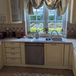 Jasna kuchnia w stylu rustykalnym z beżowymi szafkami, wzorzystą zasłoną w oknie i widokiem na ogród.