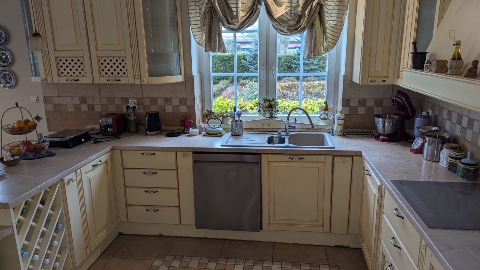 Jasna kuchnia w stylu rustykalnym z beżowymi szafkami, wzorzystą zasłoną w oknie i widokiem na ogród.