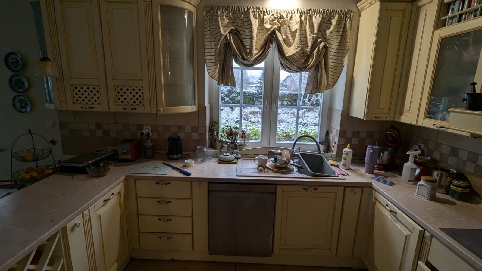 Kuchnia w stylu rustykalnym z kremowymi szafkami i oknem z widokiem na zaśnieżony ogród, naczynia w zlewie czekają na umycie, widoczne drobne zabrudzenia na blacie.