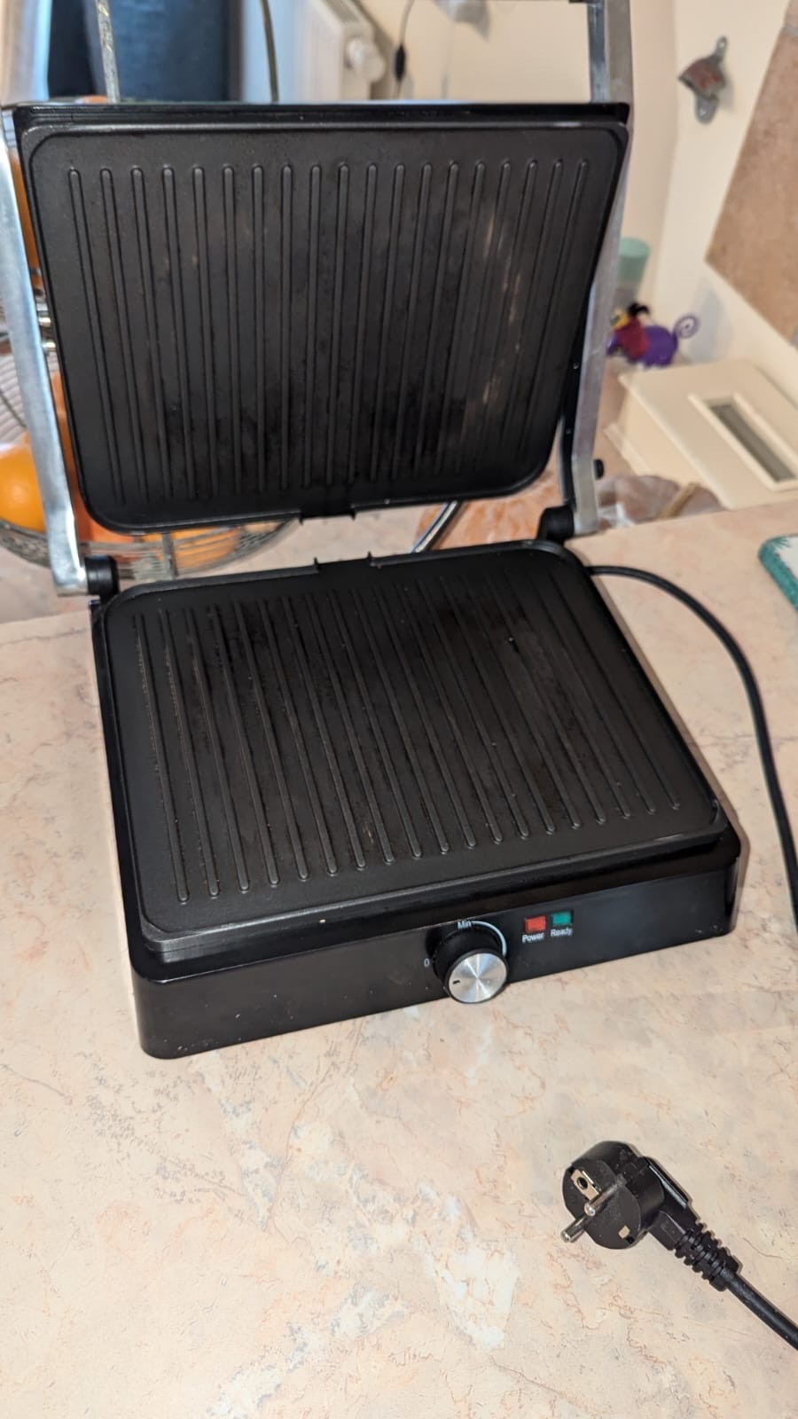 Elektryczny grill kontaktowy z otwartą górną płytą, widoczne ślady użytkowania i resztki jedzenia, kabel z wtyczką leży obok na blacie kuchennym.