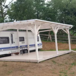 Biała drewniana pergola z płaskim dachem kryjąca przyczepę kempingową na nierównym terenie.
