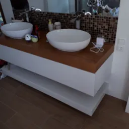 Nowoczesna łazienka z podwójną umywalką nablatową na blacie z drewna, białe szafki pod umywalką, brązowa mozaika na ścianie, widoczne akcesoria łazienkowe i zabawki.