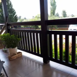 Ciemna, drewniana balustrada na tarasie z widokiem na ogród; na podłodze płytki, donica z zieloną rośliną, śpiący pies i drewniany koszyk.