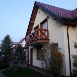 WIEWIÓR SPÓŁKA Z OGRANICZONĄ ODPOWIEDZIALNOŚCIĄ - Dwupiętrowy dom z brązową, drewnianą balustradą balkonową, widoczny z zewnątrz, ze stojącą drabiną i osobą w tle.