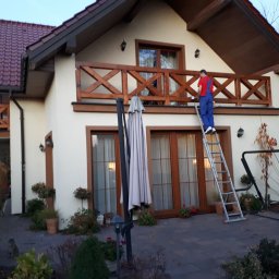 WIEWIÓR SPÓŁKA Z OGRANICZONĄ ODPOWIEDZIALNOŚCIĄ - Mężczyzna maluje drewnianą balustradę balkonu domu jednorodzinnego, stojąc na drabinie; widoczna elewacja, okna z drewnianymi ramami i otoczenie domu.