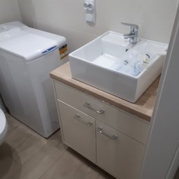 WIEWIÓR SPÓŁKA Z OGRANICZONĄ ODPOWIEDZIALNOŚCIĄ - Nowoczesna łazienka z białą umywalką nablatową, szafką pod umywalką z beżowym blatem i pralką ładowaną od góry, widoczna instalacja elektryczna