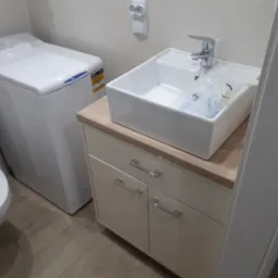 Nowoczesna łazienka z białą umywalką nablatową, szafką pod umywalką z beżowym blatem i pralką ładowaną od góry, widoczna instalacja elektryczna