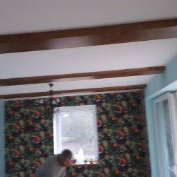 WIEWIÓR SPÓŁKA Z OGRANICZONĄ ODPOWIEDZIALNOŚCIĄ - Wnętrze pokoju z zamontowanymi dekoracyjnymi belkami stropowymi z drewna, widoczny fragment osoby pracującej przy oknie