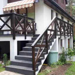 Zewnętrzne schody z ciemnoszarego kompozytu prowadzące na taras z ciemnobrązdką balustradą krzyżową, widok z dołu.