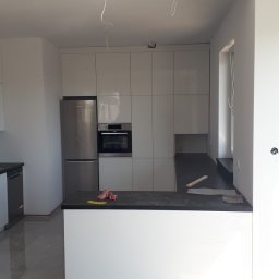 WIEWIÓR SPÓŁKA Z OGRANICZONĄ ODPOWIEDZIALNOŚCIĄ - Nowoczesna, jasna kuchnia z białymi szafkami, czarnym blatem i srebrnymi urządzeniami w trakcie montażu, widoczne wystające kable i niedokończone ściany.