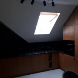 Kuchnia w skosach z drewnianymi frontami, czarnym blatem i oknem dachowym, oświetlona reflektorami punktowymi.