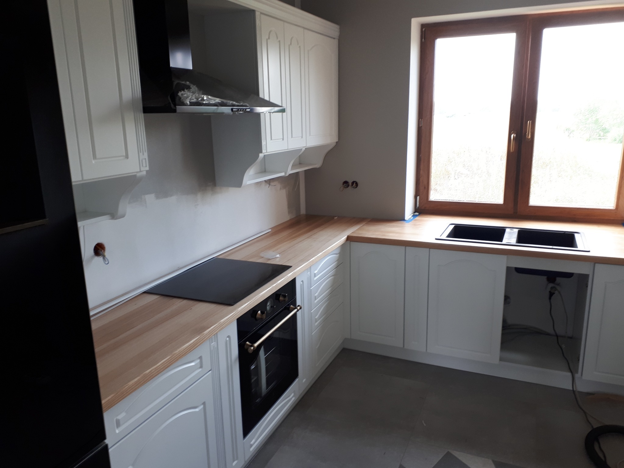 Nowoczesna kuchnia w trakcie montażu: białe szafki z frezowanymi frontami, drewniany blat, czarna płyta indukcyjna i piekarnik z mosiężnymi detalami, widoczne niedokończone prace wykończeniowe...