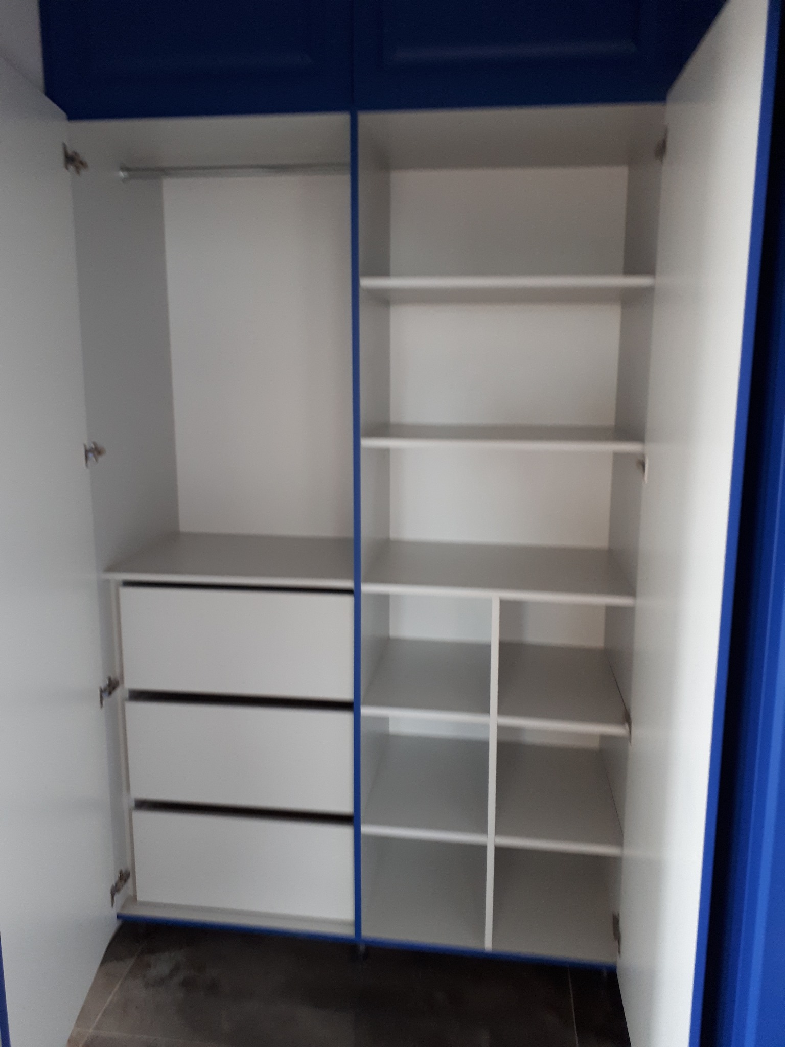 Wnętrze szafy wnękowej z otwartymi drzwiami, widoczne trzy szuflady po lewej stronie i półki po prawej, białe elementy z niebieskimi akcentami.