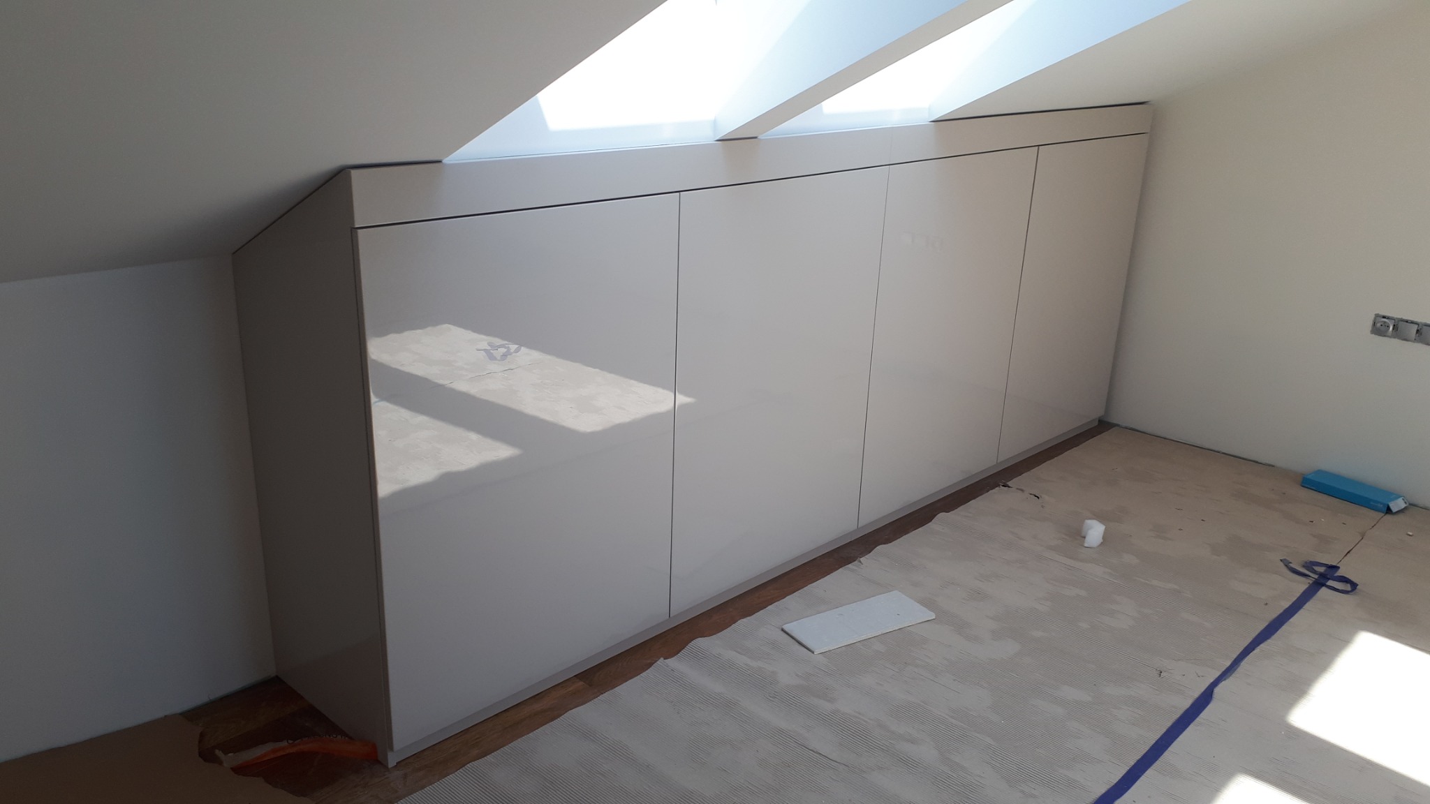Biała szafa na wymiar z frontami na wysoki połysk, zabudowana pod skosem dachu, widoczna podczas montażu na podłodze z resztkami kleju i oznaczeniami.