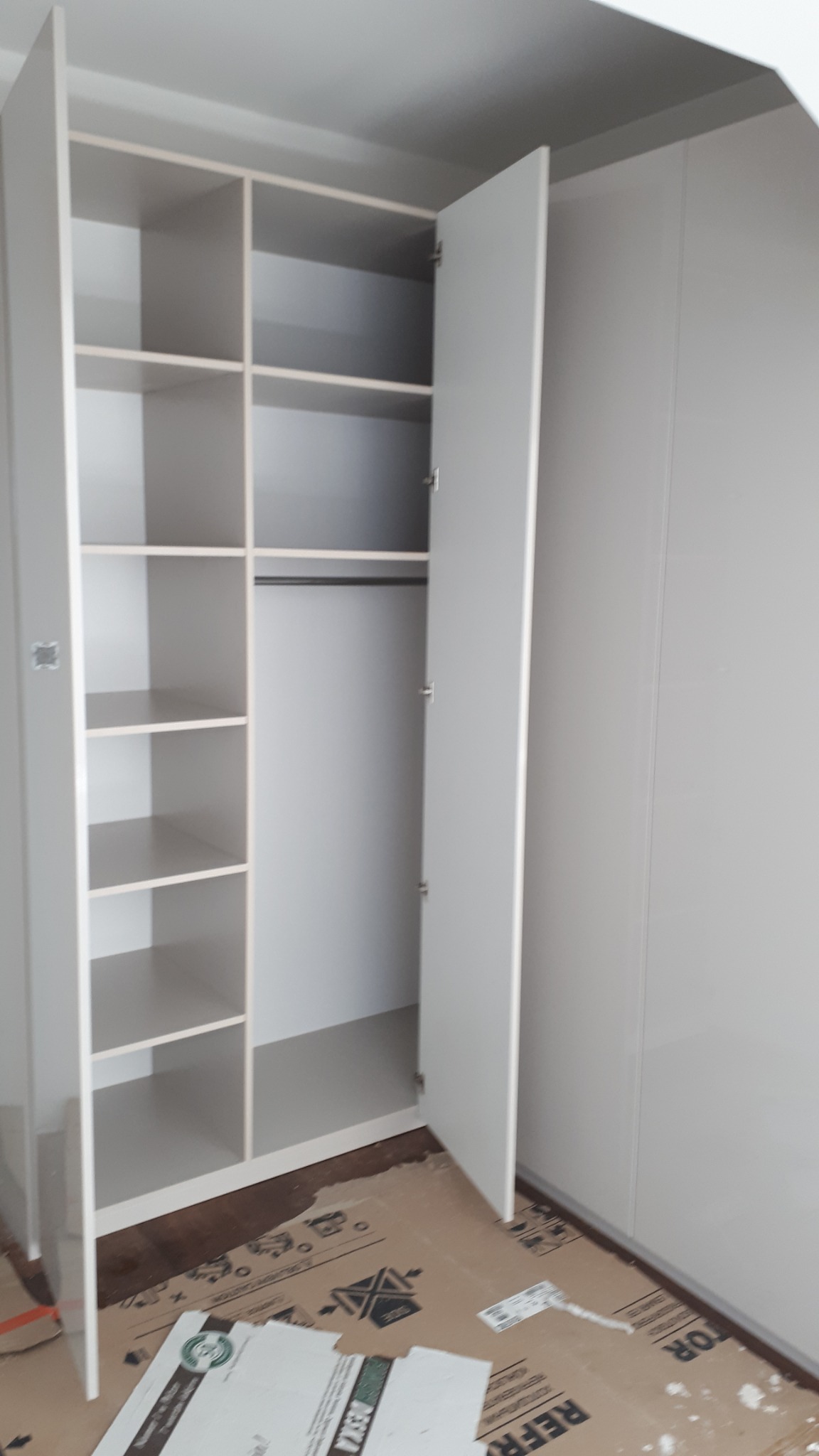 Wnętrze szafy wnękowej z otwartymi drzwiami, widoczne półki i drążek na ubrania, podłoga zabezpieczona kartonem podczas montażu.